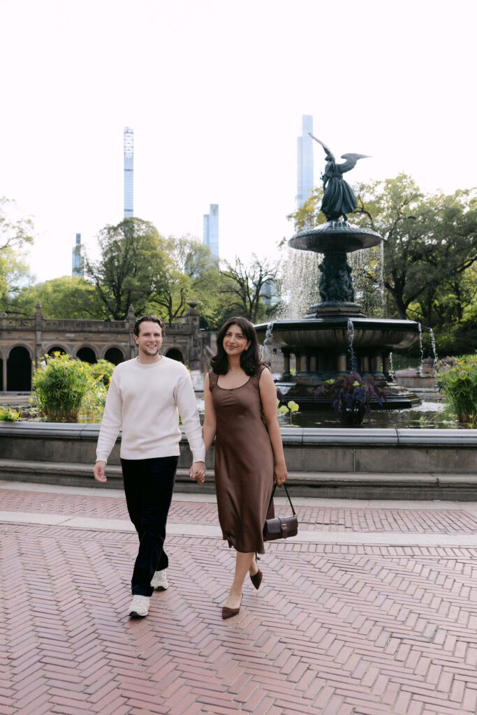 nyc engagement session photo of a couple walking through central park near a fountain