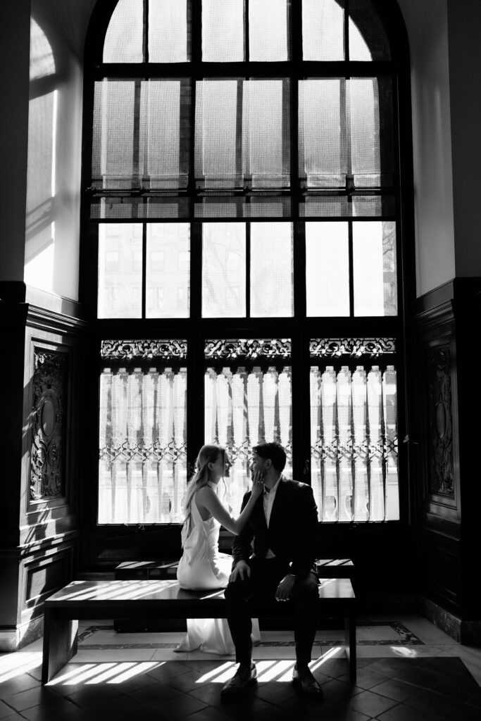 Black and white photo of couple by large window inside New York Public Library during NYC engagement session
