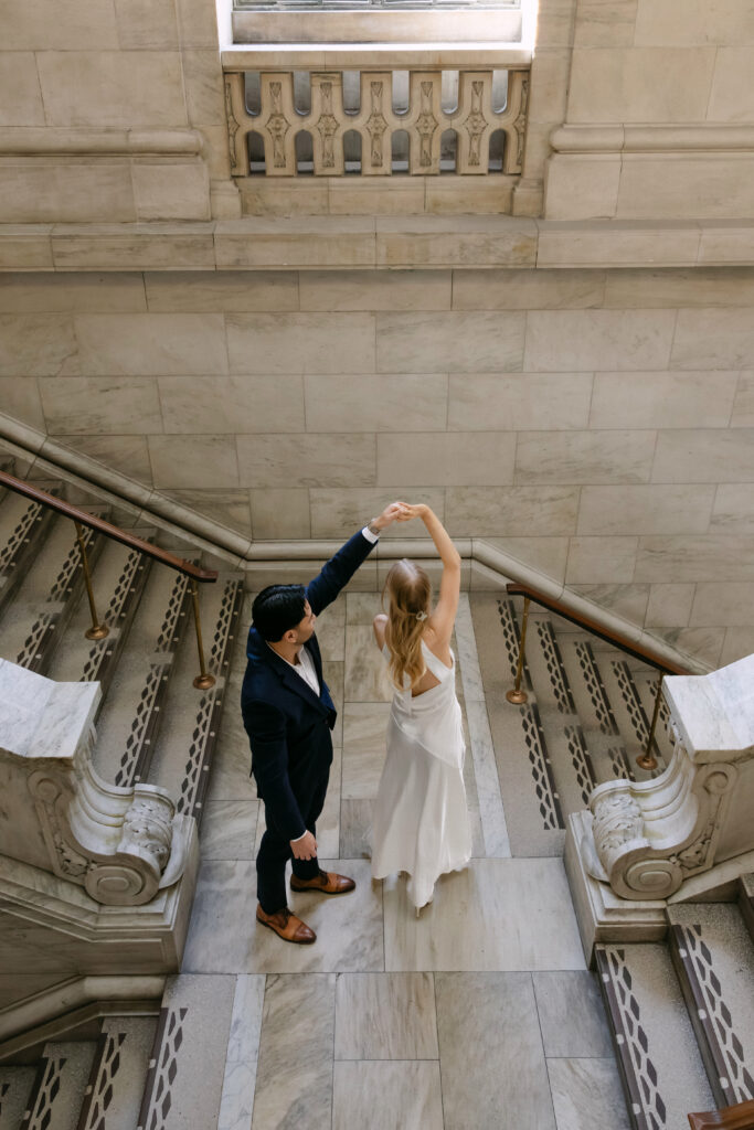 Couple twirling on marble staircase inside New York Public Library during romantic NYC engagement session