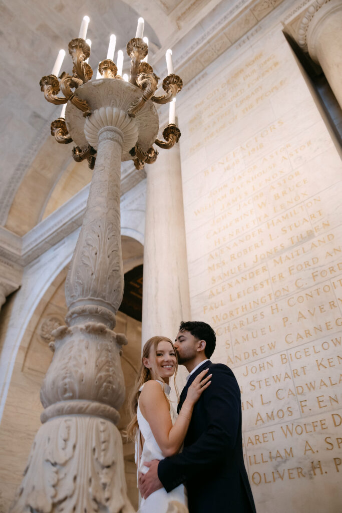 Couple embracing beneath chandelier inside New York Public Library during NYC engagement photos