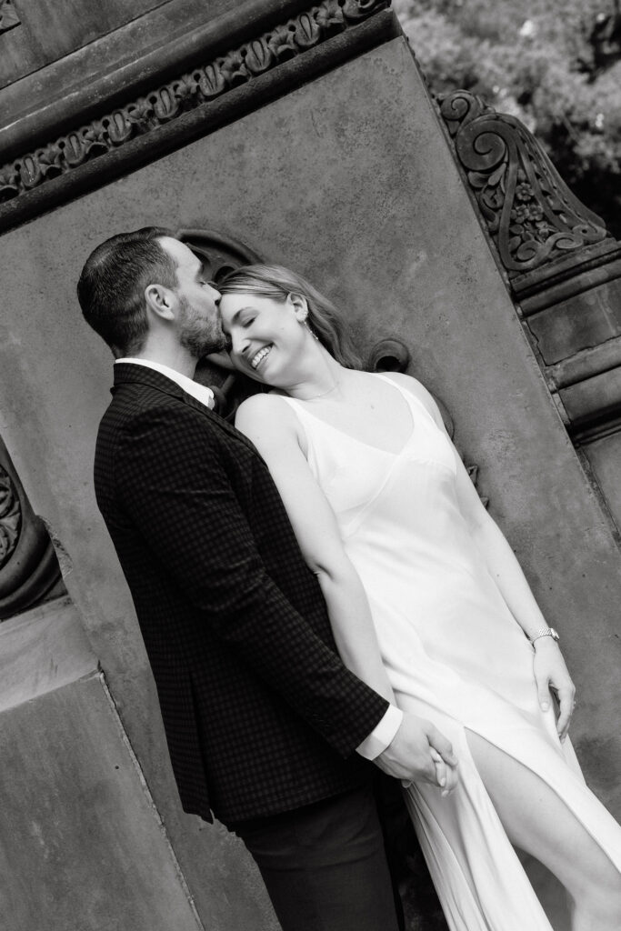 Black-and-white portrait of an engaged couple embracing beside the stone architecture at Bethesda Terrace in Central Park.