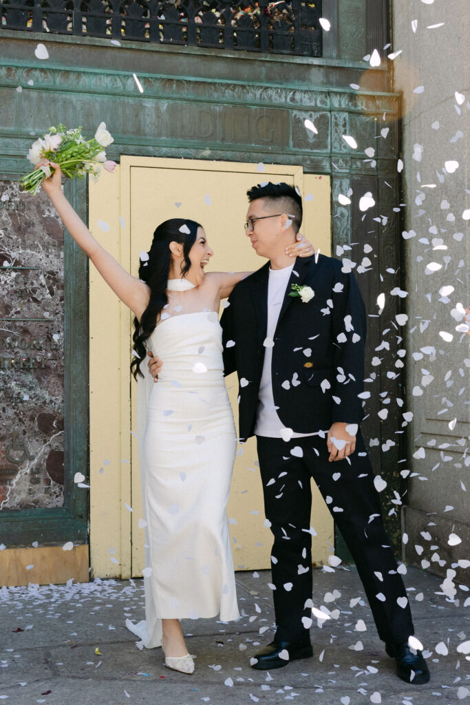 Newlyweds celebrating under white confetti outside the Manhattan Marriage Bureau after their City Hall wedding.