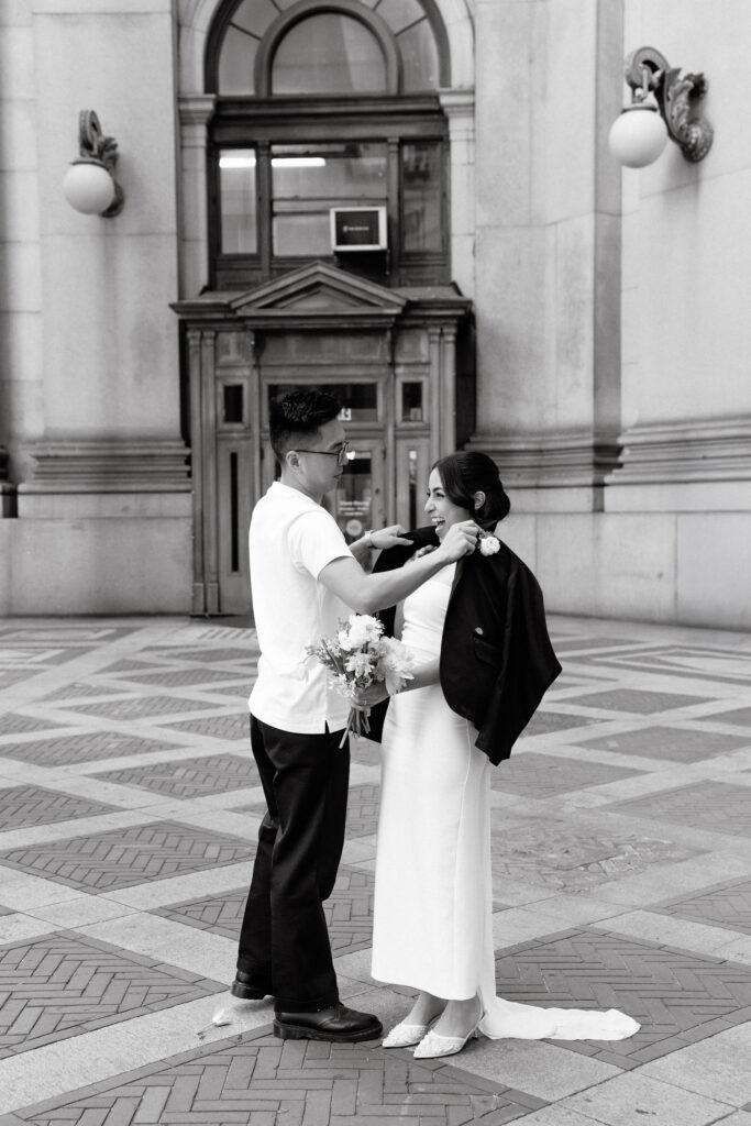Groom helping the bride put on his jacket during their Manhattan City Hall wedding portraits.
