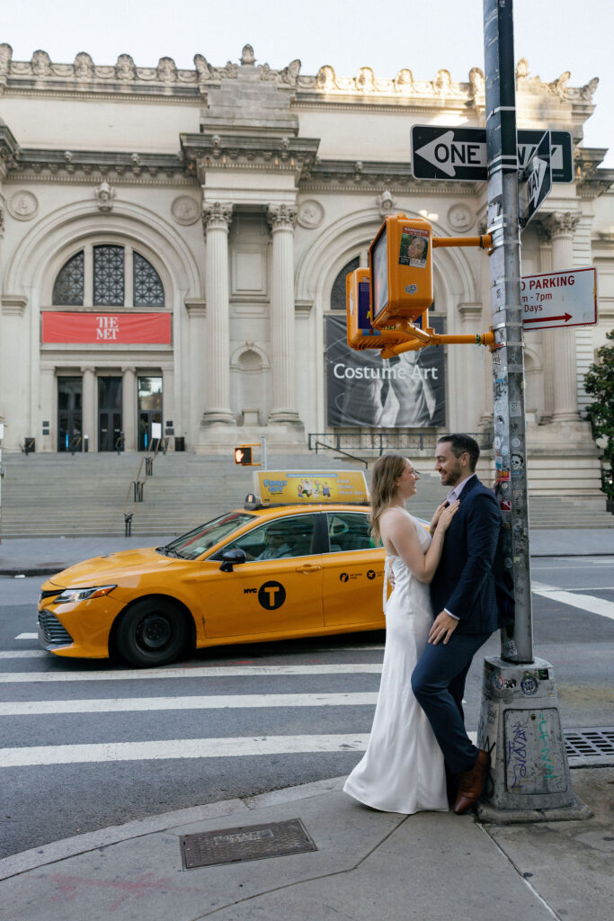 Engaged couple posing beside a yellow taxi with The Met in the background during their Upper East Side engagement session.