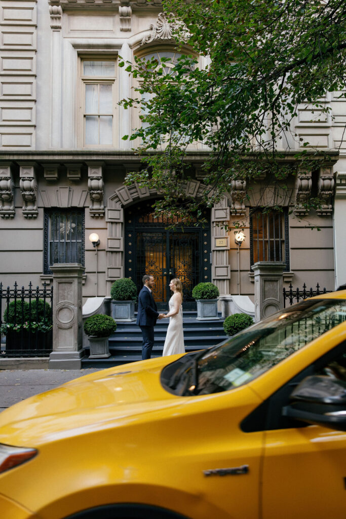 Engaged couple holding hands outside an elegant Upper East Side townhouse as a yellow taxi passes in the foreground.
