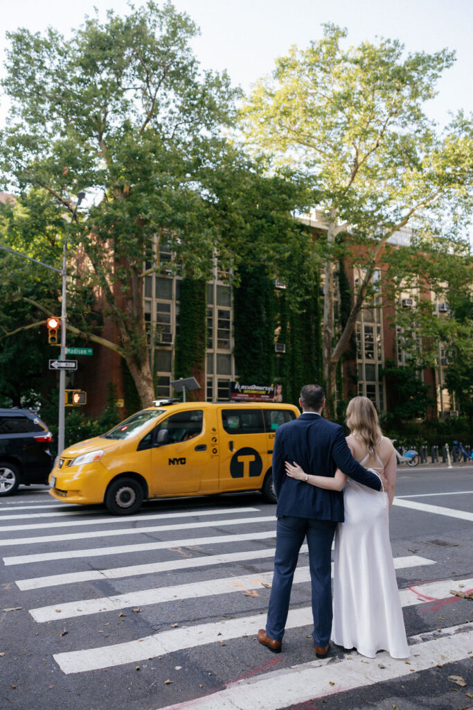 Engaged couple embracing in an Upper East Side crosswalk with a yellow taxi passing behind them.